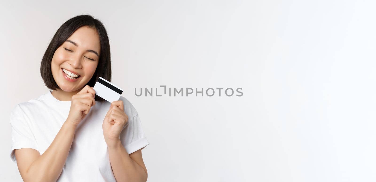 Image of smiling asian woman hugging credit card, buying contactless, standing in white tshirt over white background by Benzoix