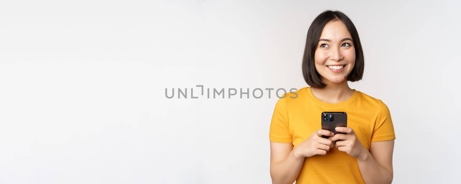 People and technology concept. Smiling asian girl using smartphone, texting on mobile phone, standing against white background.