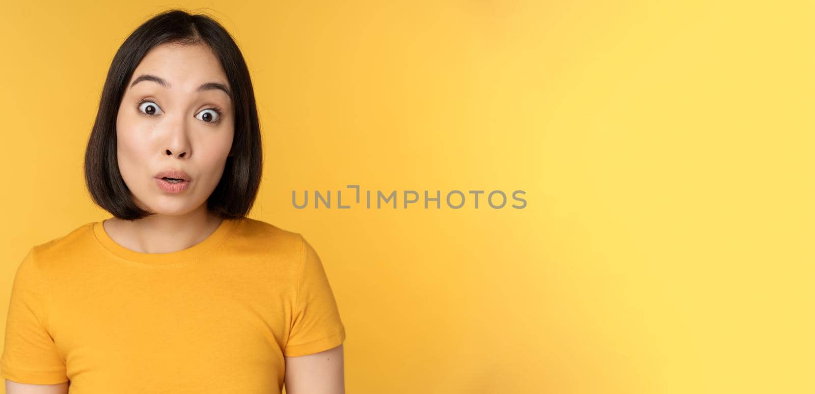 Close up portrait of asian girl showing surprised reaction, raising eyebrows amazed, reacting to big news, standing over yellow background by Benzoix