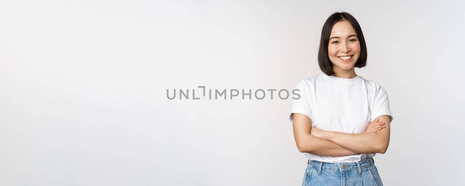 Portrait of happy asian woman smiling, posing confident, cross arms on chest, standing against studio background by Benzoix