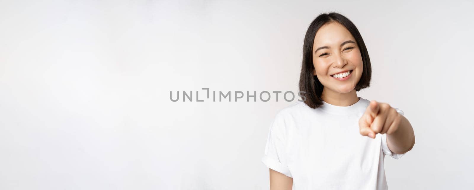 Portrait of happy smiling asian woman with white teeth, pointing finger at camera, choosing you, congratulating, standing over white background.