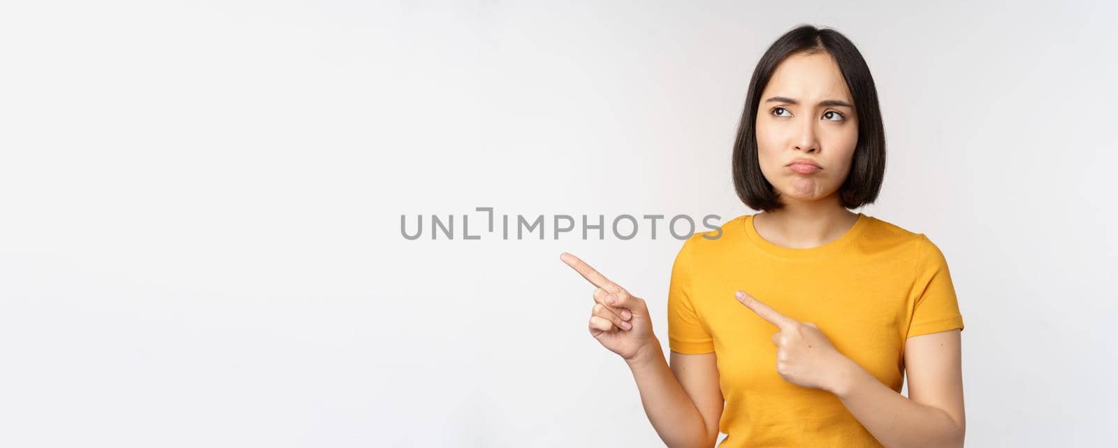 Upset grumpy asian girl, pouting and looking at smth unfair, pointing fingers left at banner, logo brand, standing in yellow tshirt over white background by Benzoix
