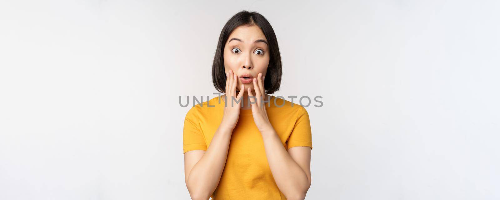 Close up portrait of asian woman looking surprised, wow face, staring impressed at camera, standing over white background in yellow t-shirt.