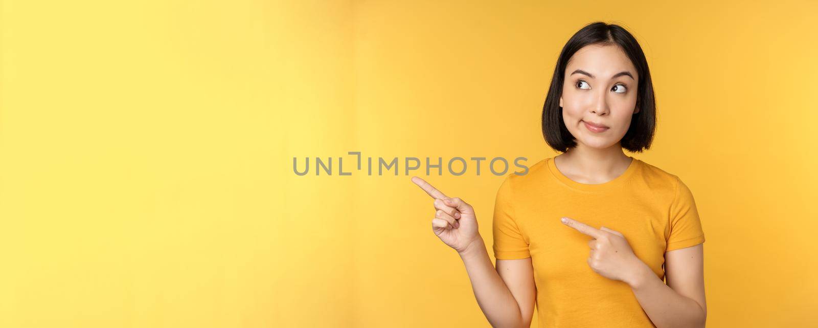 Portrait of intrigued asian woman, looking and pointing fingers left at advertisement, showing smth interesting, standing over yellow background.