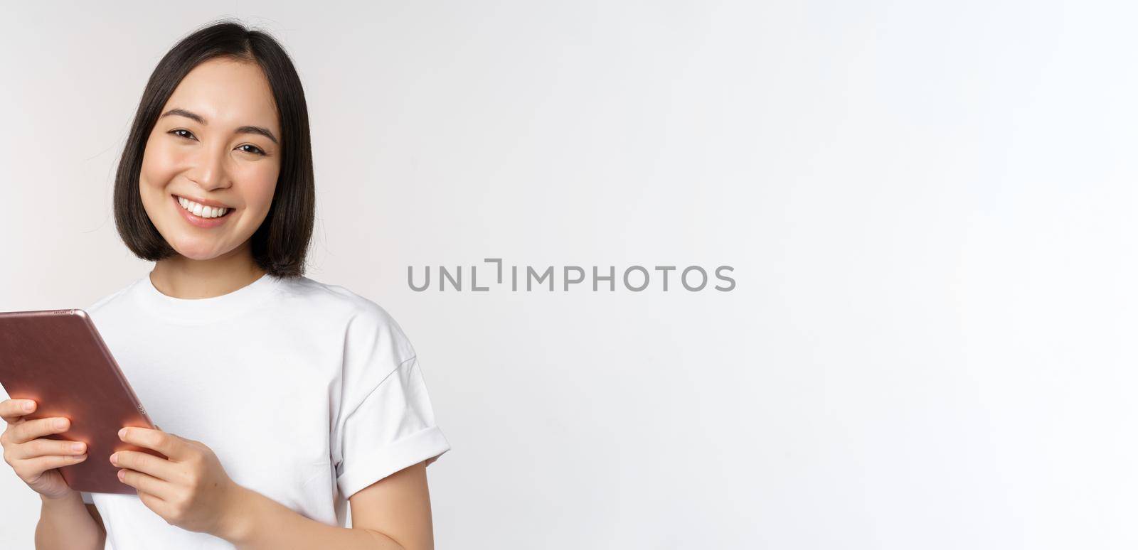 Smiling asian girl with digital tablet, looking happy and laughing, posing in tshirt over white background. Copy space