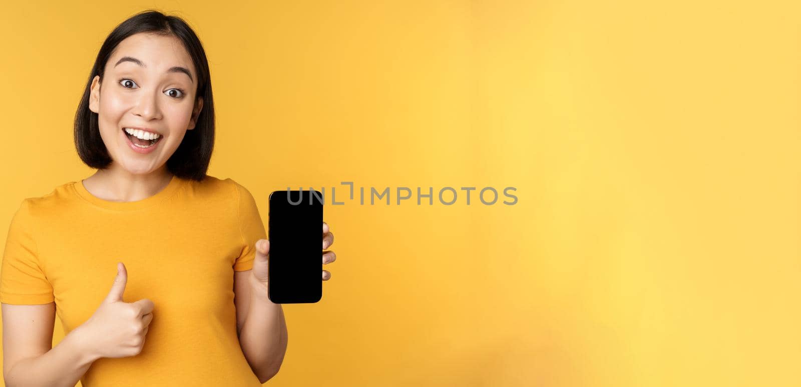 Excited asian woman showing mobile phone screen, thumbs up, like smth good, recommending smartphone application, standing over yellow background by Benzoix