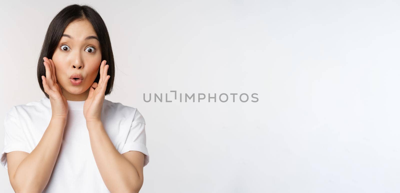 Close up portrait of asian girl looking surprised, wow face, reacting amazed at smth, standing in white tshirt over studio background, isolated by Benzoix