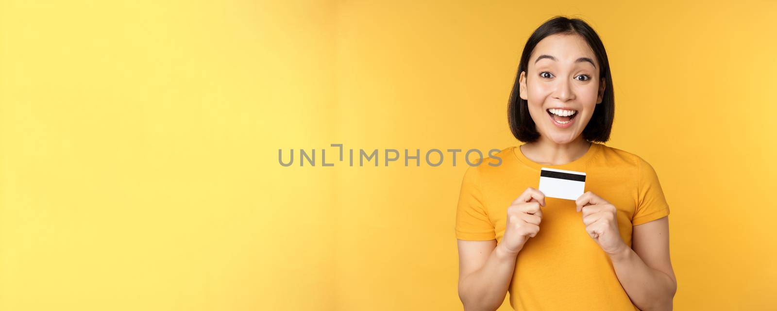 Beautiful asian woman showing credit card and smiling, recommending bank service, standing over yellow background by Benzoix