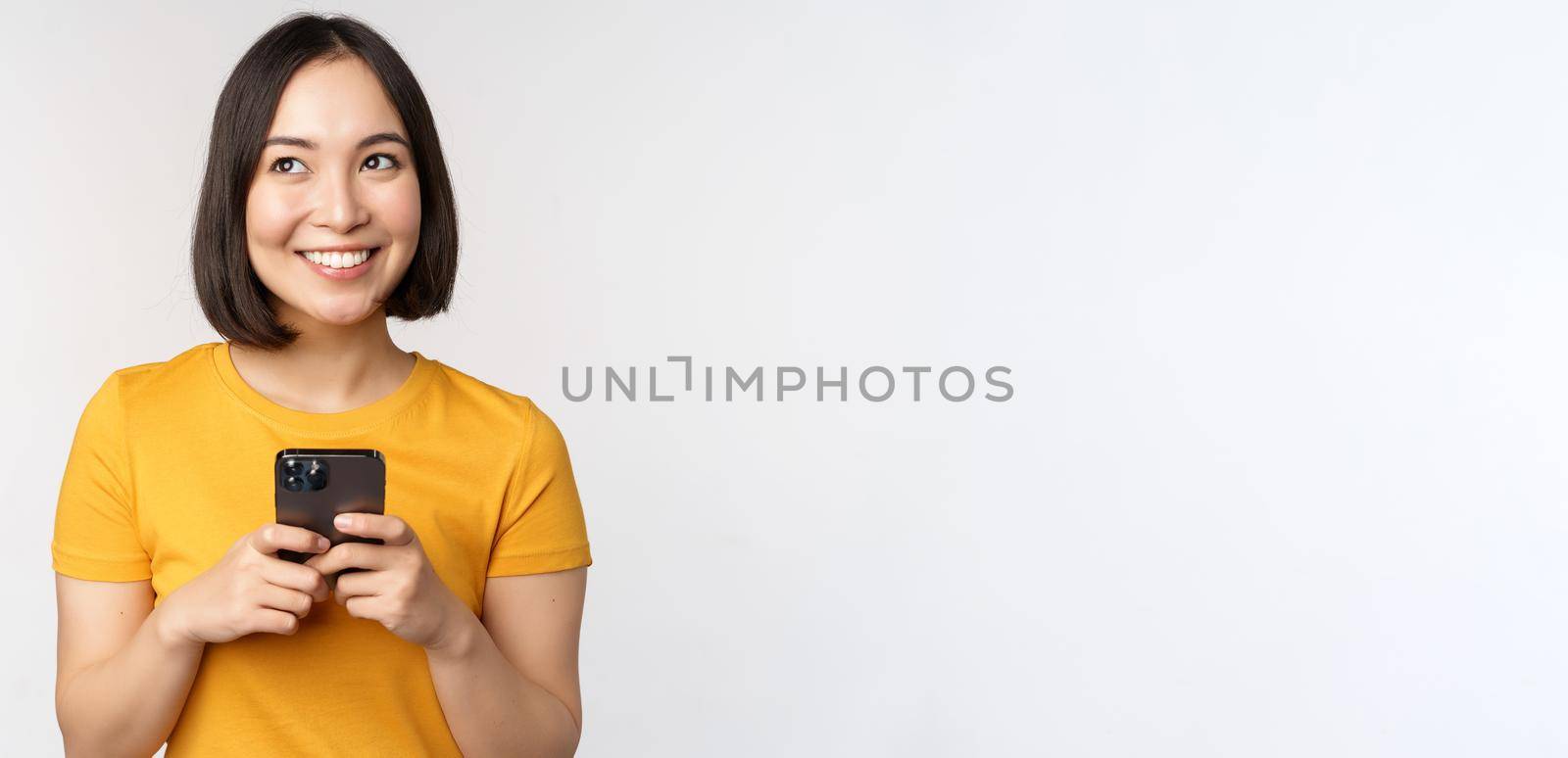 People and technology concept. Smiling asian girl using smartphone, texting on mobile phone, standing against white background.