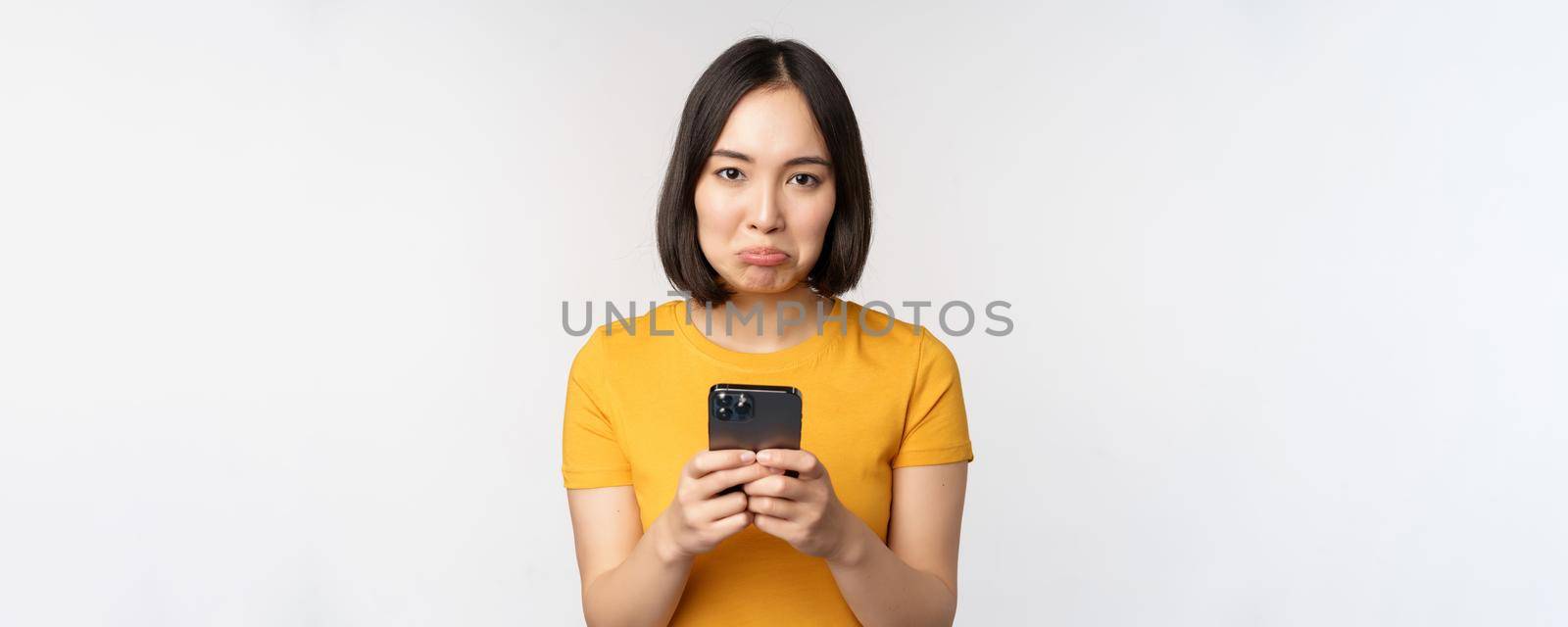 Sad asian woman holding smartphone, looking upset with regret, standing in yellow tshirt against white background.