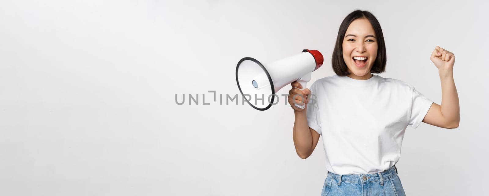 Happy asian woman shouting at megaphone, making announcement, advertising something, standing over white background.