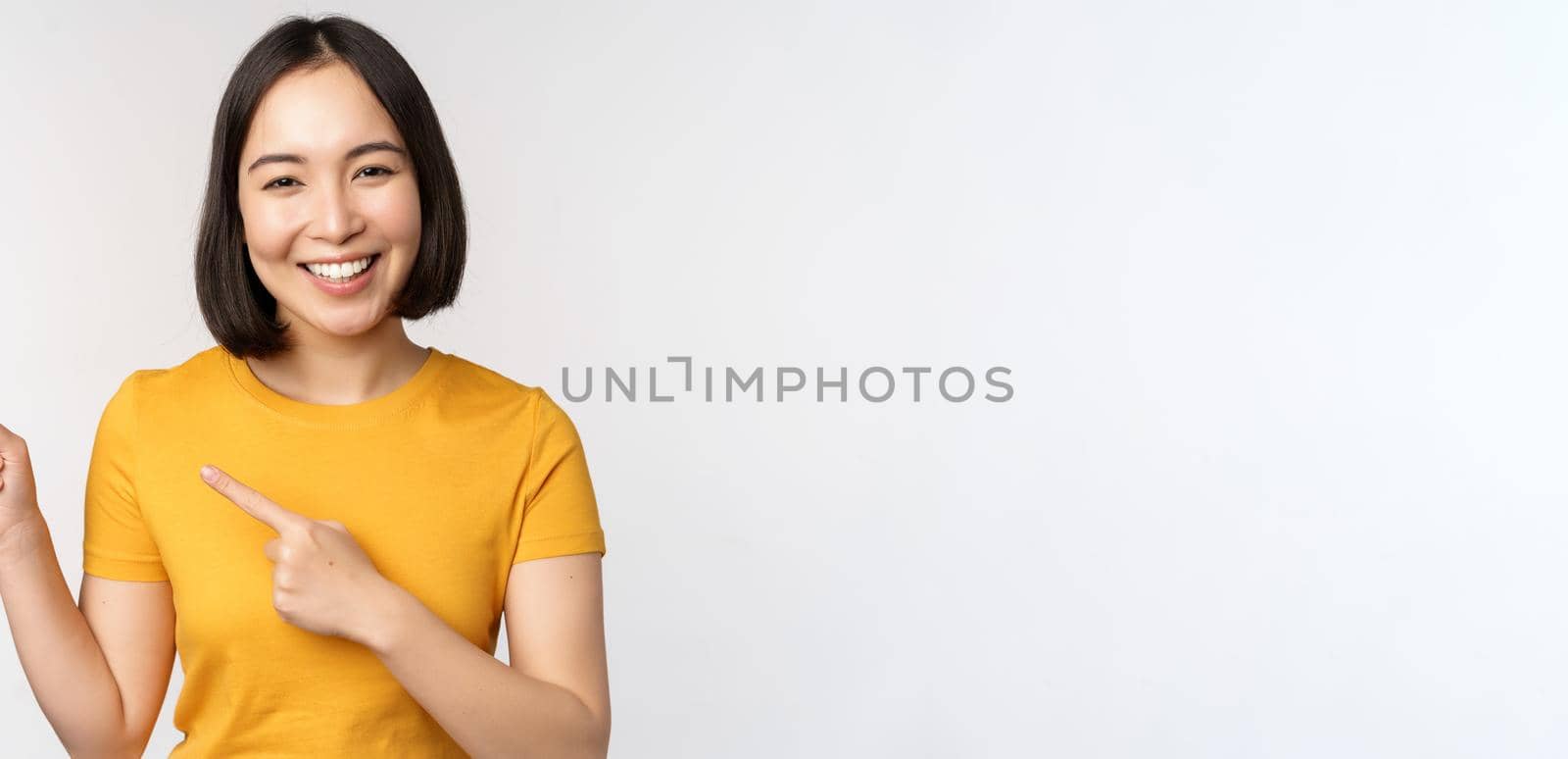 Portrait of smiling asian brunette girl in yellow tshirt, pointing fingers left, showing copy space, promo deal, demonstrating banner, standing over white background.