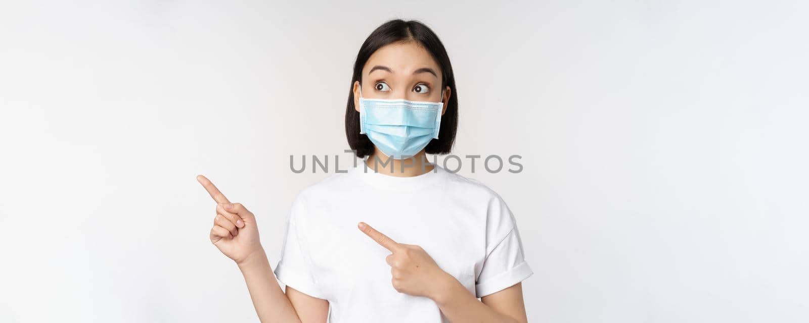 Young korean woman in medical face mask pointing fingers left and looking at logo, showing advertisement or banner, standing over white background.