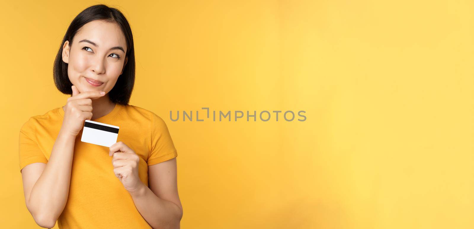 Thinking asian woman holding credit card and smiling, planning buy smth, standing thoughtful over yellow background.