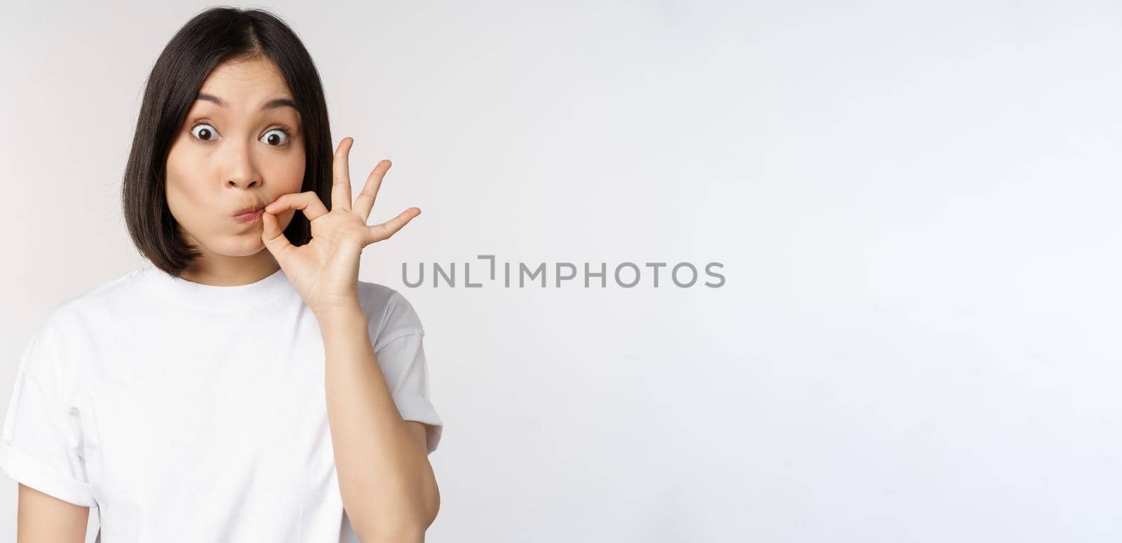 Image of young asian woman showing silence gesture, zipping mouth, seal lips, make promise, standing over white background.