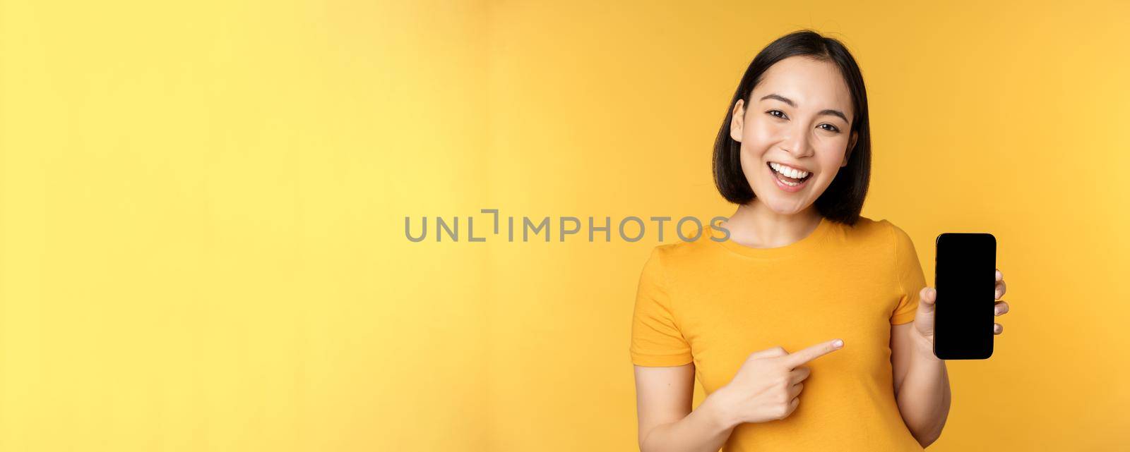 Beautiful happy asian girl showing mobile phone screen, application on smartphone gadget, standing over yellow background.