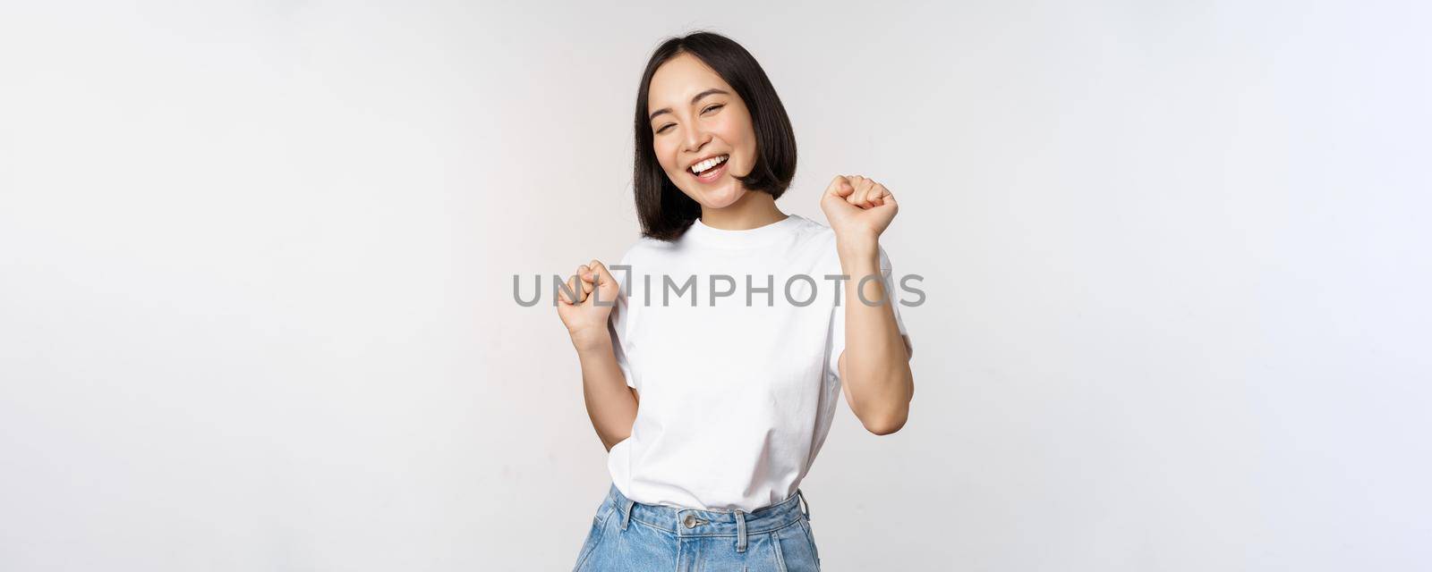 Happy dancing korean girl posing against white background, wearing tshirt.