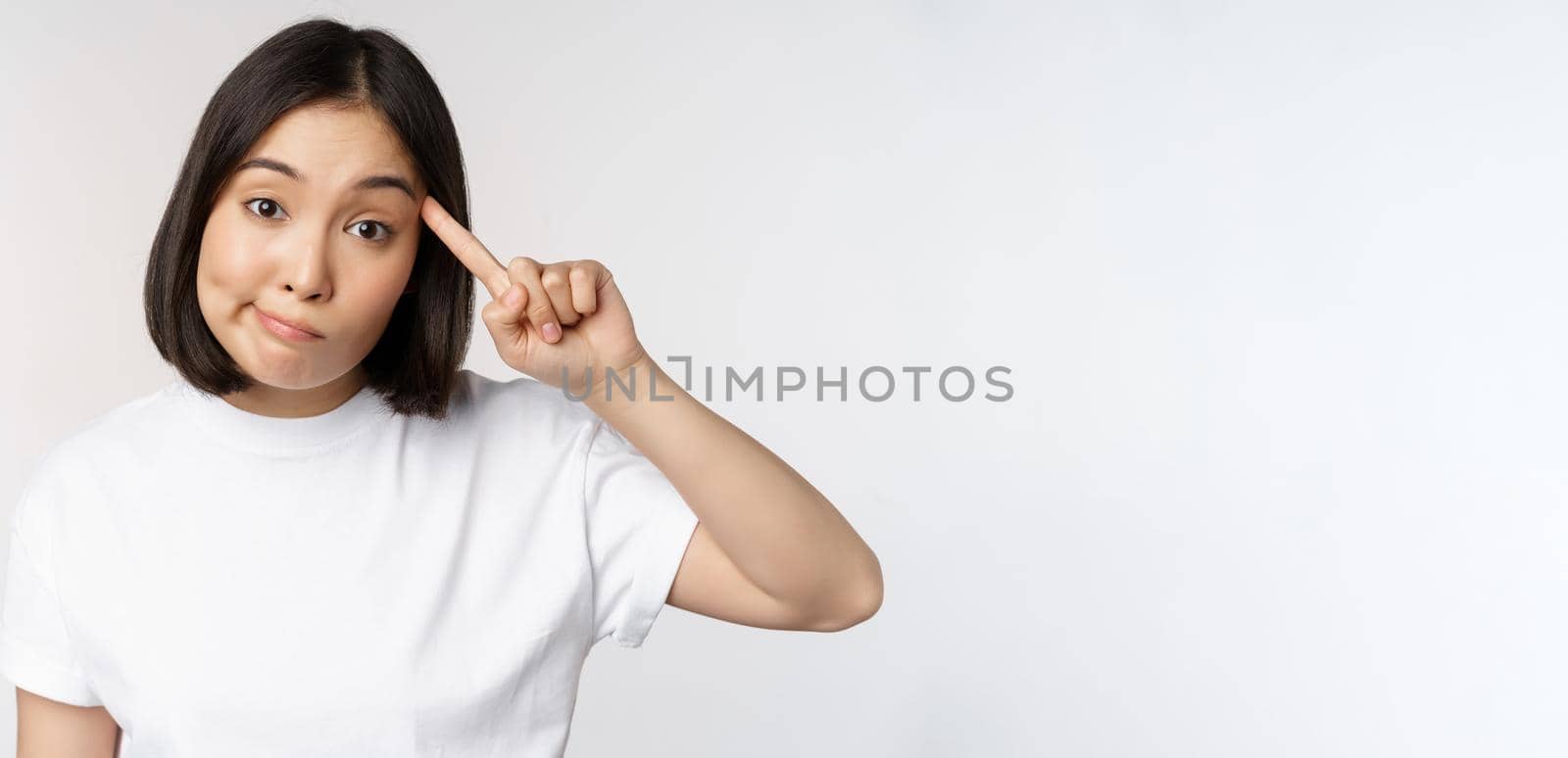 Are you crazy. Young asian woman roll finger on head, mocking someone stupid, standing in tshirt over white background. Copy space