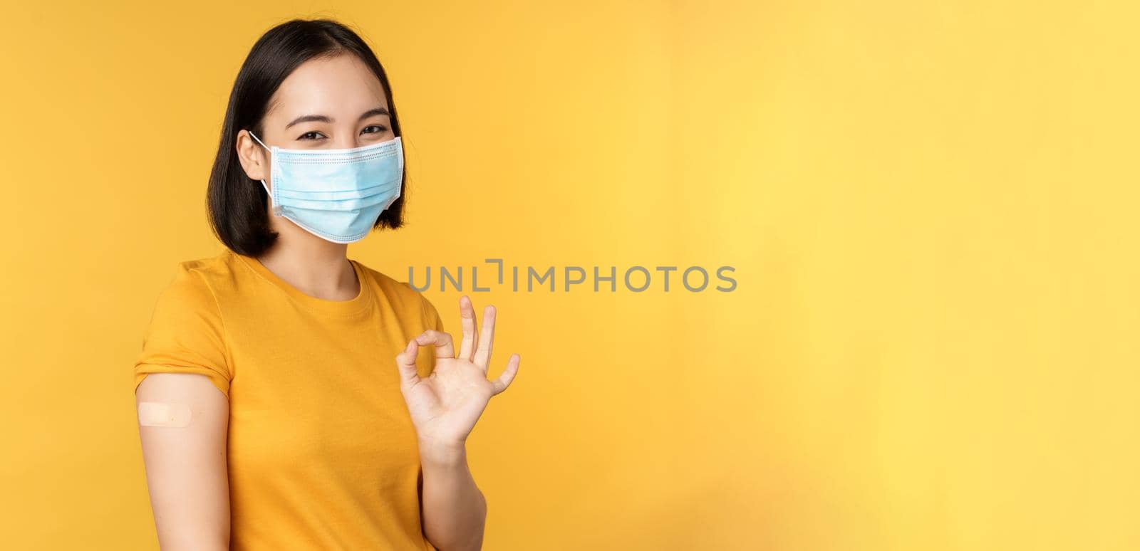 Vaccination from covid and health concept. Happy asian girl showing okay, wearing medical mask, band aid on shoulder, got coronavirus vaccine shot, yellow background.