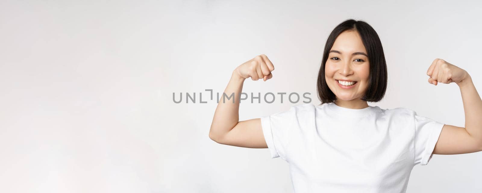 Smiling asian woman showing flexing biceps, muscles strong arms gesture, standing in white tshirt over white background.