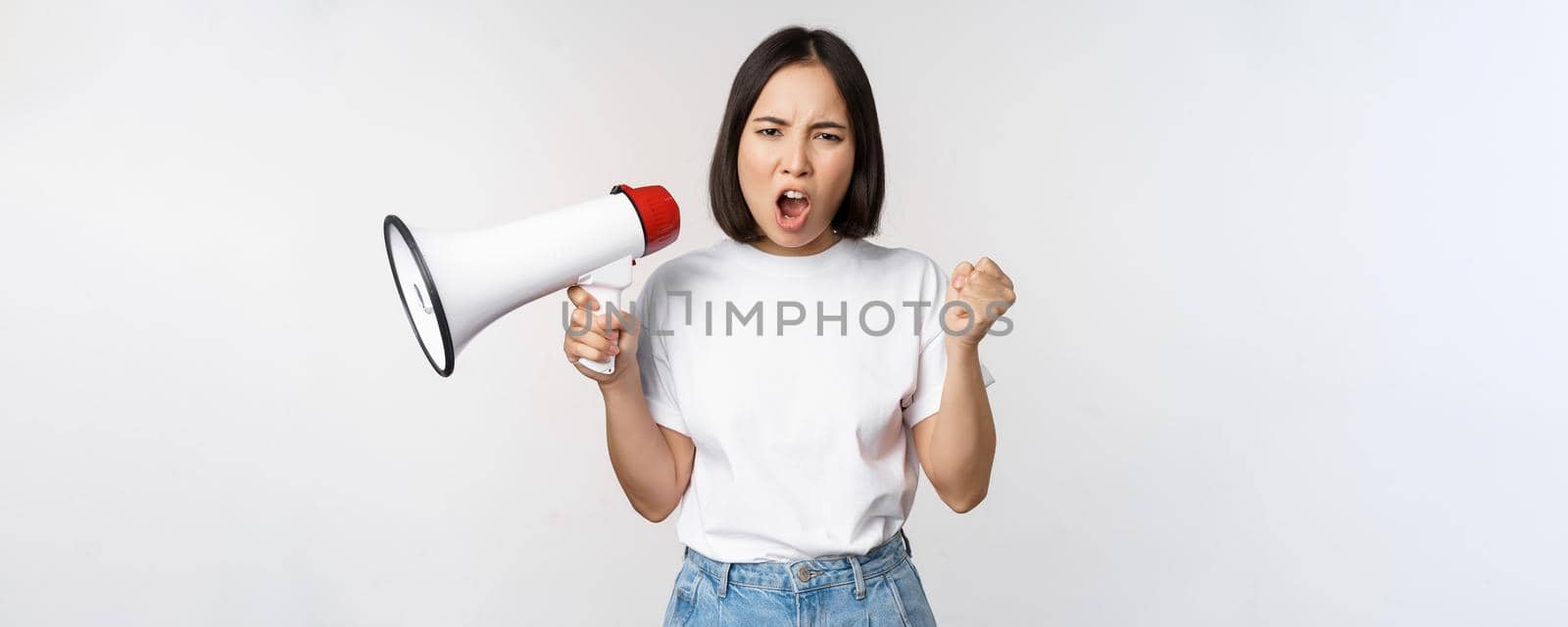 Angry asian girl activist, holding megaphone and looking furious, protesting, standing over white background. Copy space