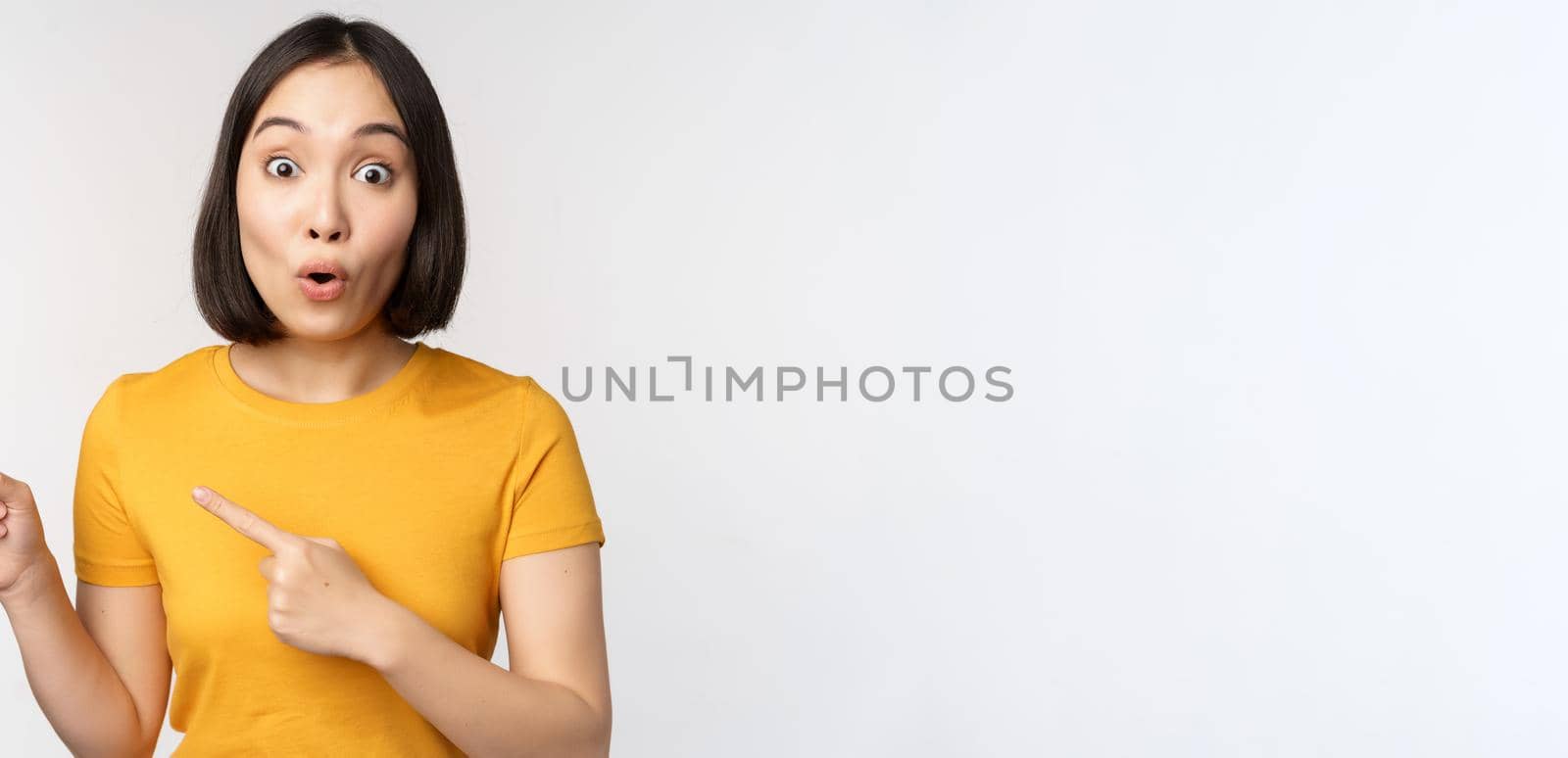 Portrait of smiling asian brunette girl in yellow tshirt, pointing fingers left, showing copy space, promo deal, demonstrating banner, standing over white background.