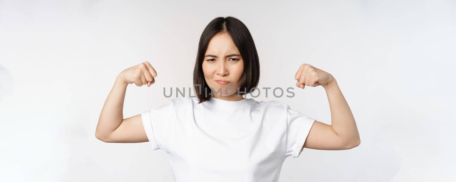 Smiling asian woman showing flexing biceps, muscles strong arms gesture, standing in white tshirt over white background.