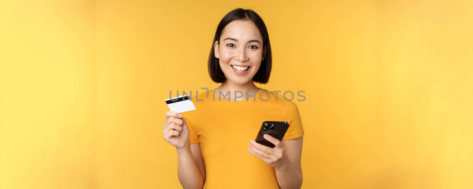 Online shopping. Cheerful asian girl holding credit card and smartphone, paying, order with mobile phone, standing over yellow background.