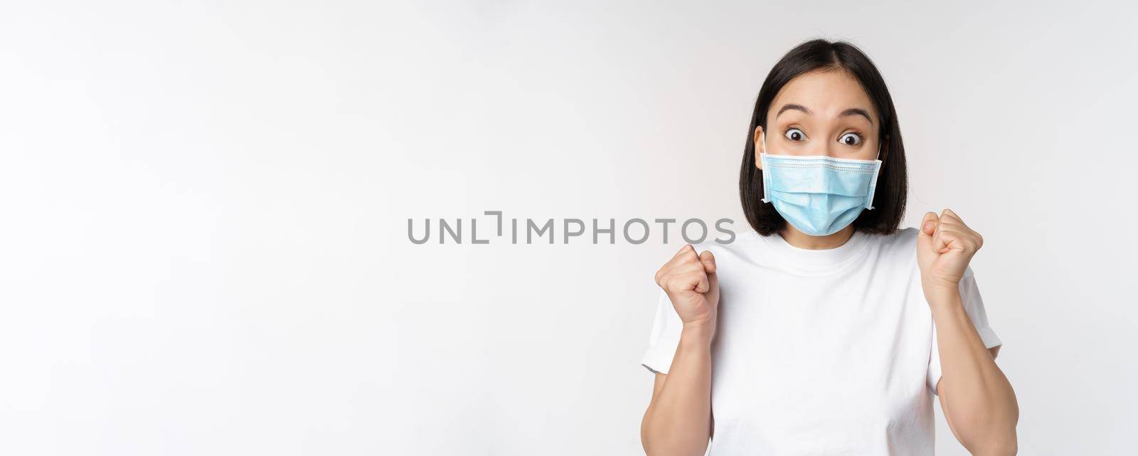 Covid-19, healthcare and medical concept. Enthusiastic asian woman in medical face mask, dancing and celebrating, winning, achieve goal, standing over white background by Benzoix