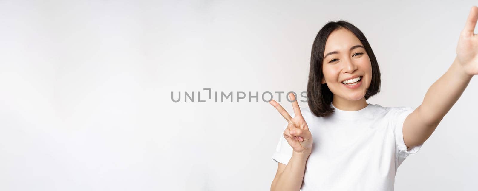 Beautiful young asian woman taking selfie, posing with peace v-sign, smiling happy, take photo, posing against white background.