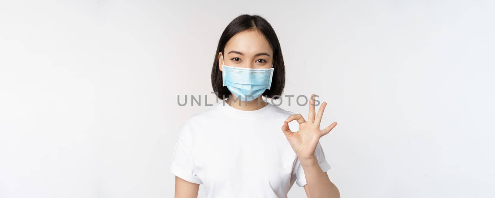Covid-19, healthcare and medical concept. Happy asian girl smiling, wearing medical face mask, showing okay, ok sign, approve, recommending smth, white background.