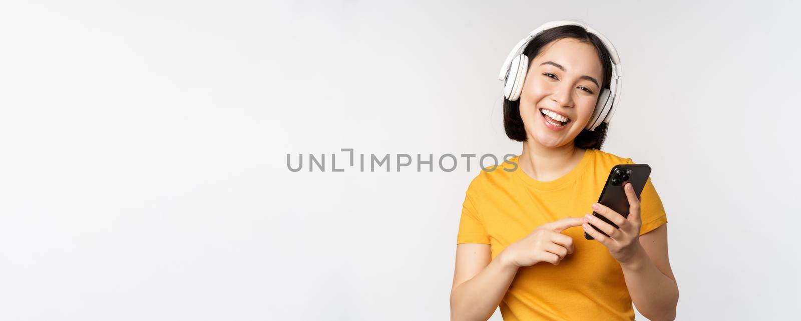 Cute japanese girl in headphones, looking at mobile phone and smiling, using music app on smartphone, standing against white background.