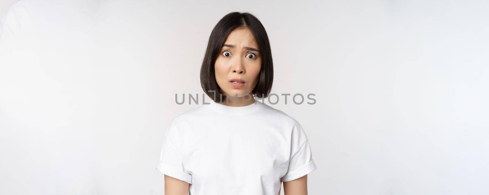 Image of shocked and frustrated asian woman, looking alarmed at camera, cant understand, puzzled, standing over white background.
