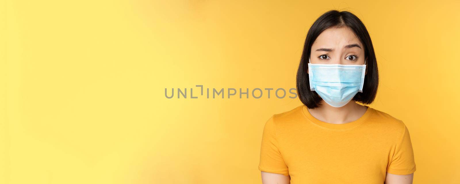 Portrait of skeptical and confused asian woman in medical face mask, raising eyebrow doubtful, standing over yellow background by Benzoix