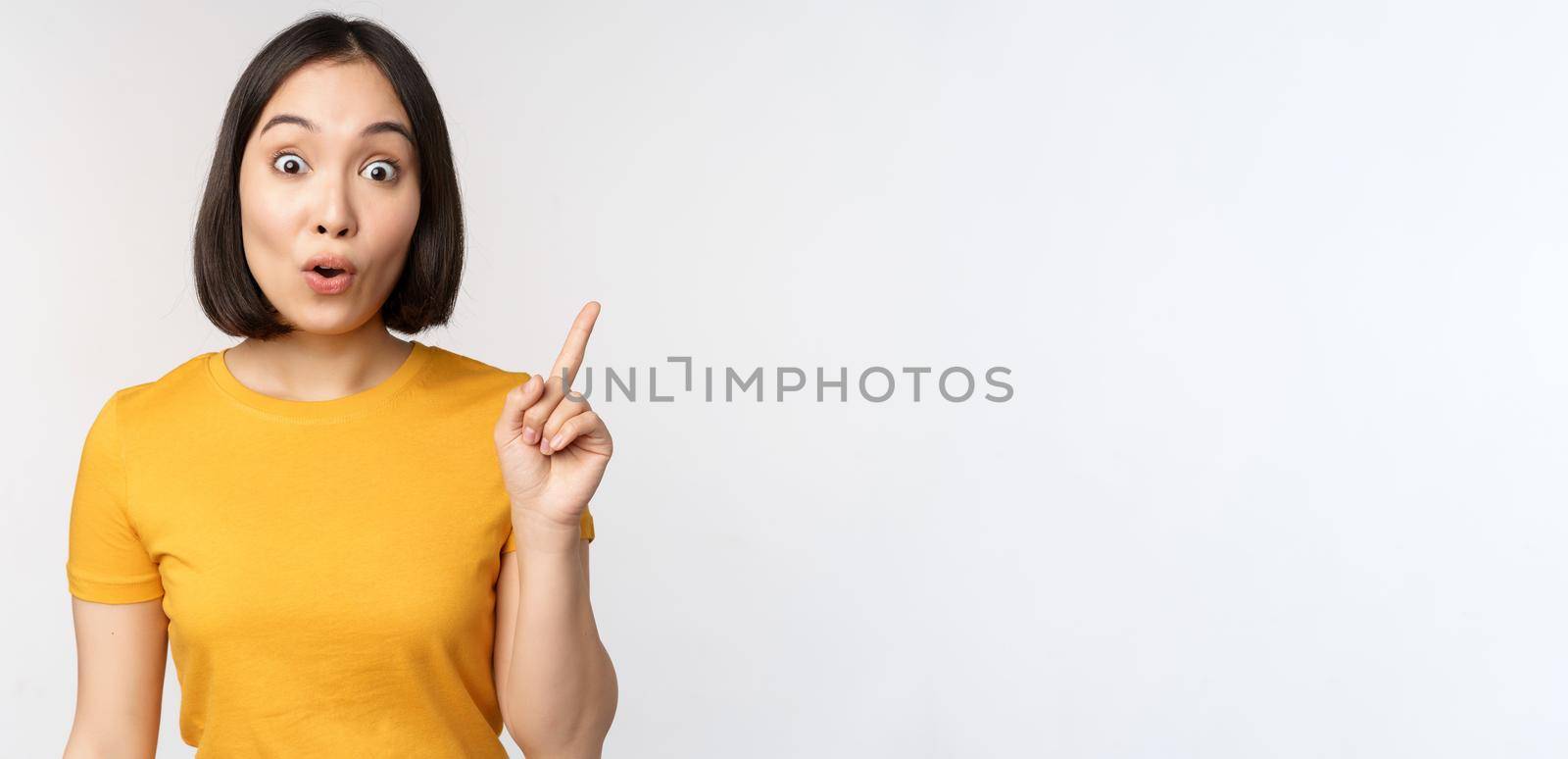 Beautiful young asian woman pointing finger up, smiling and looking amused at camera, showing advertisement, announcement on top, white background.