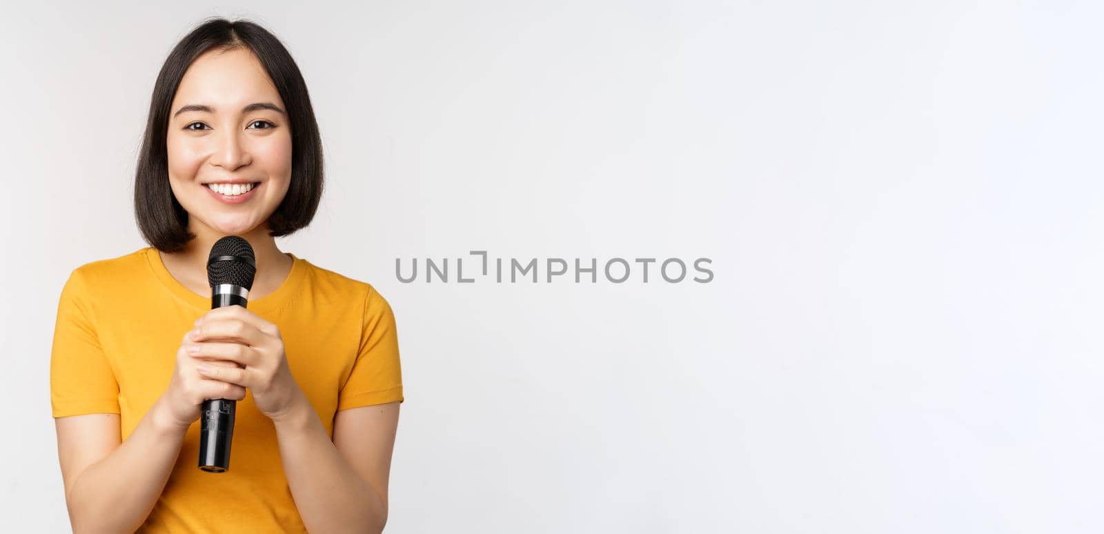 Image of young asian woman talking in microphone, perfom with mic, giving speech, standing in yellow tshirt against white background by Benzoix