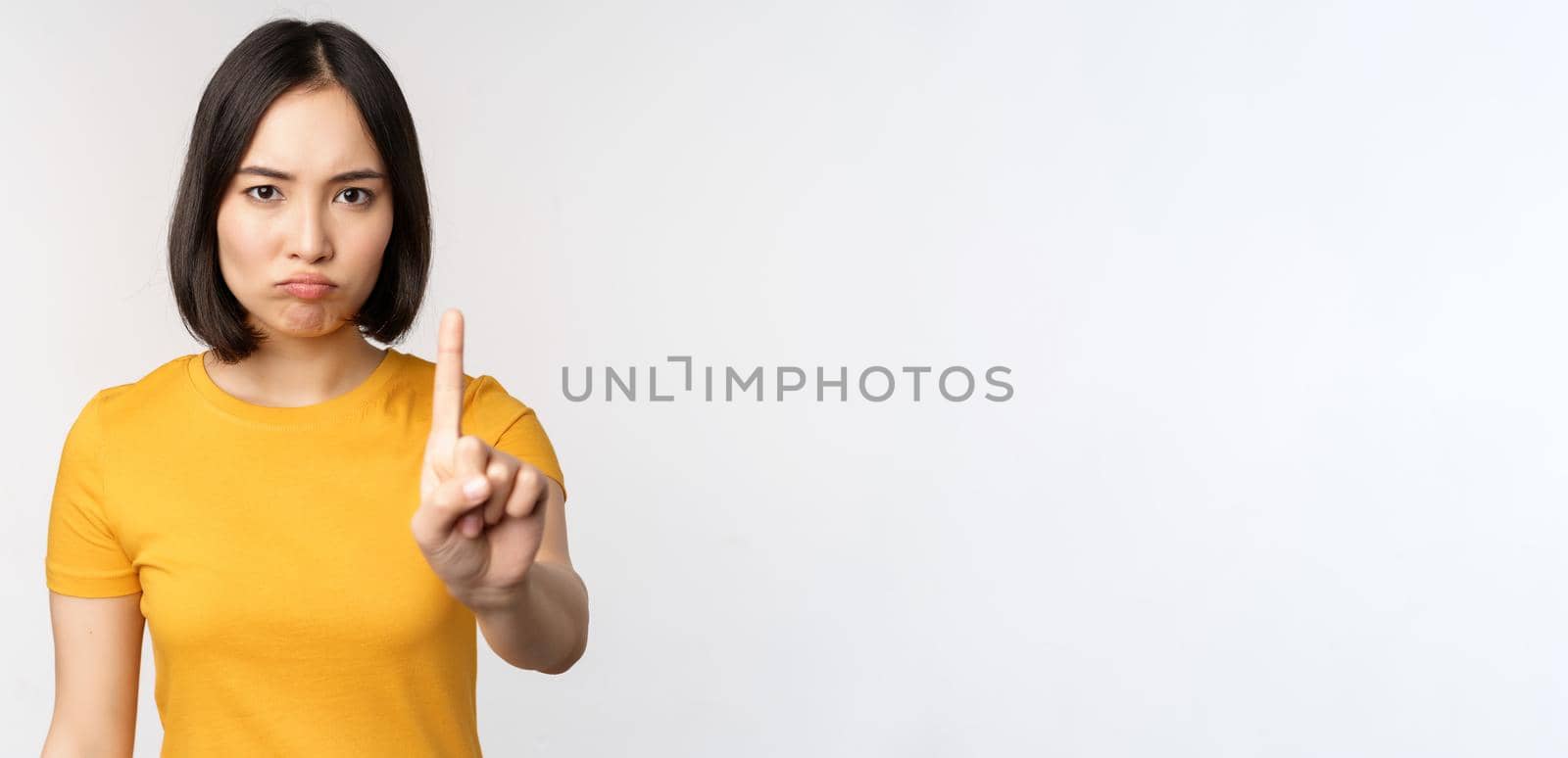 Portrait of asian woman looking serious and angry, showing stop prohibit gesture, taboo sign, forbidding smth, standing in yellow tshirt over white background by Benzoix