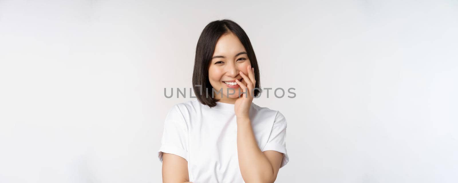 Portrait of young beautiful woman, korean girl laughing and smiling, looking coquettish, standing against white background.