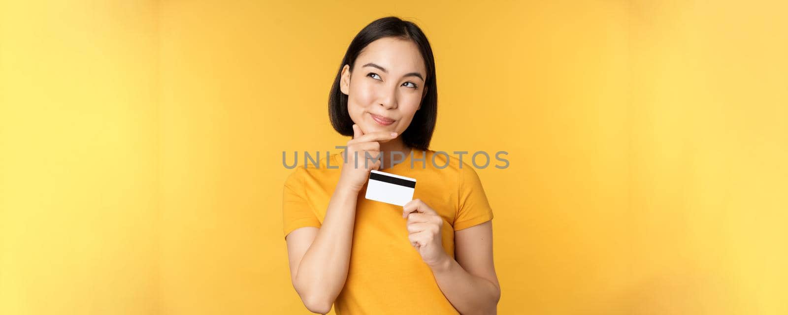 Thinking asian woman holding credit card and smiling, planning buy smth, standing thoughtful over yellow background by Benzoix