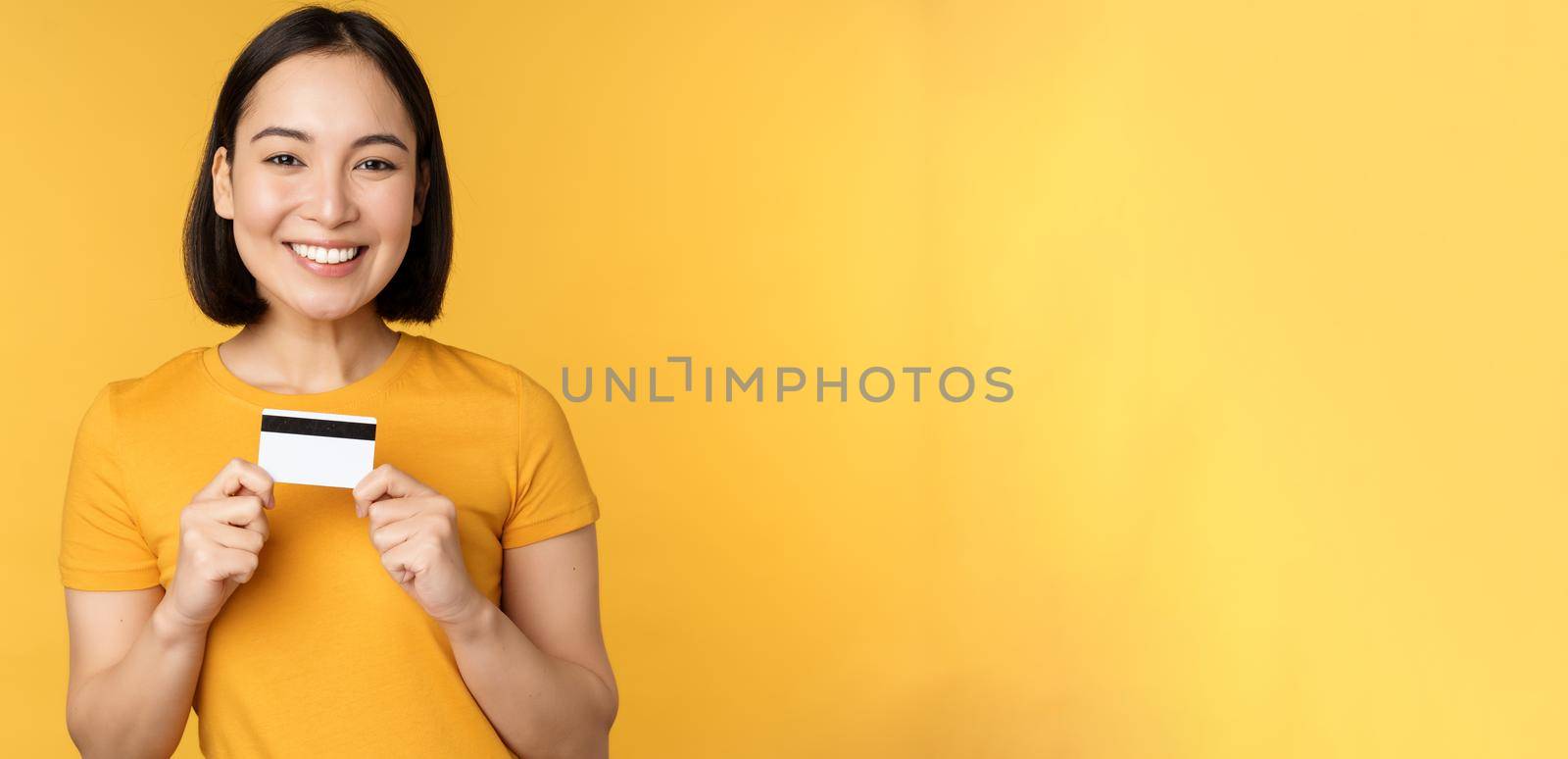 Beautiful asian woman showing credit card and smiling, recommending bank service, standing over yellow background. Copy space