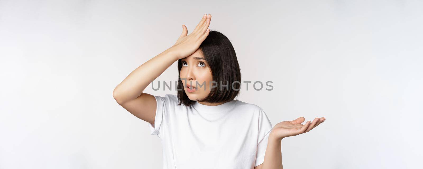 Annoyed korean girl facepalm, slap forehead and shrugging, confused by smth, standing over white background.