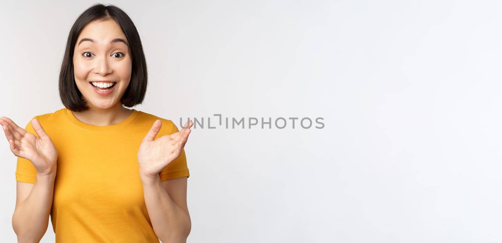 Close up portrait of asian woman looking surprised, wow face, staring impressed at camera, standing over white background in yellow t-shirt.