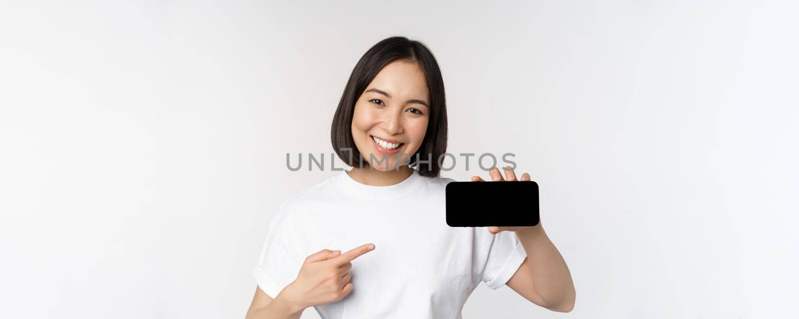 Portrait of smiling korean woman pointing finger at mobile phone screen, showing horizontal smartphone display, recommending website or store online, white background by Benzoix
