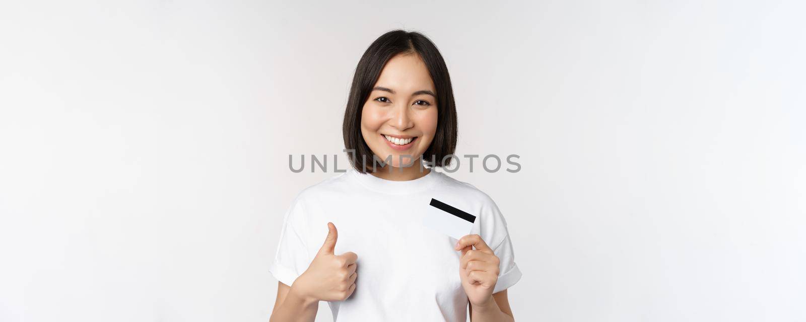 Portrait of beautiful young modern asian woman, showing credit card and thumbs up, recommending contactless payment, standing over white background.