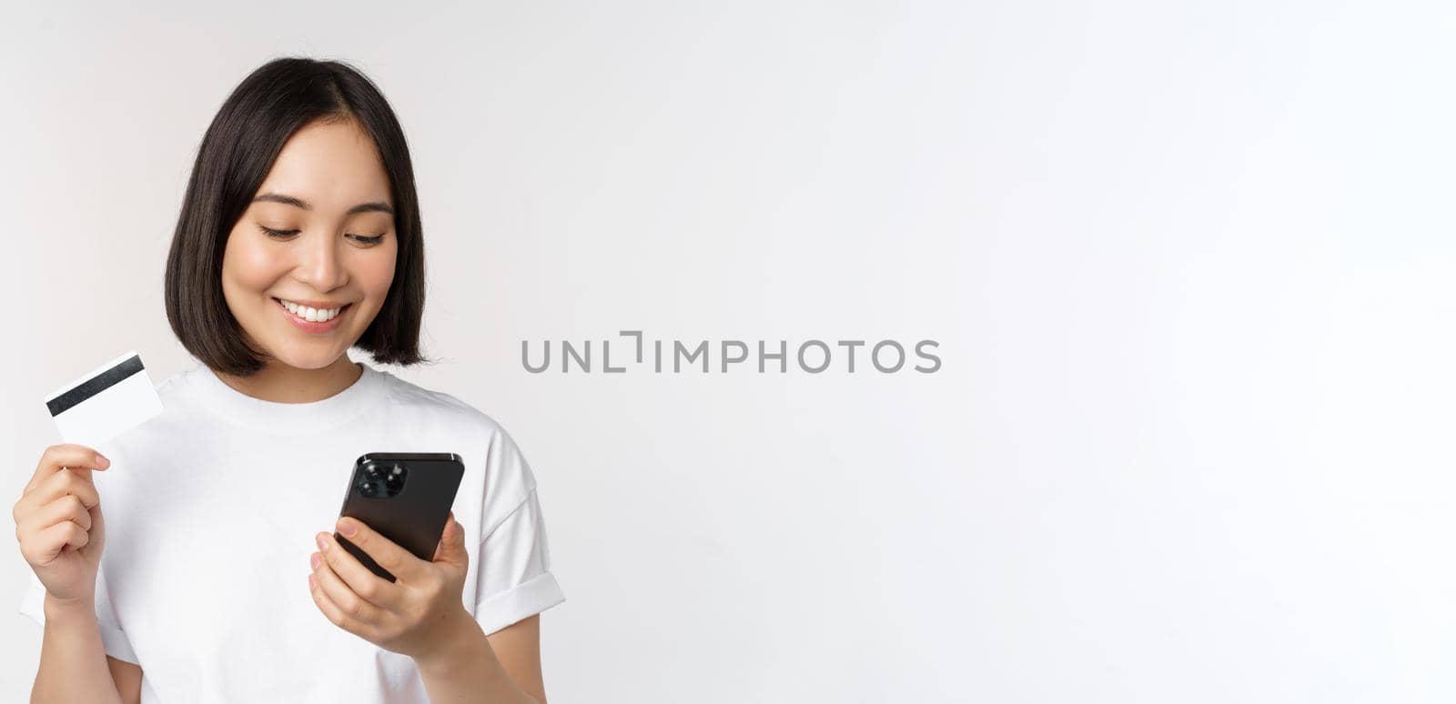 Online shopping. Happy asian woman using credit card and smartphone app, paying on website via mobile phone, white background.