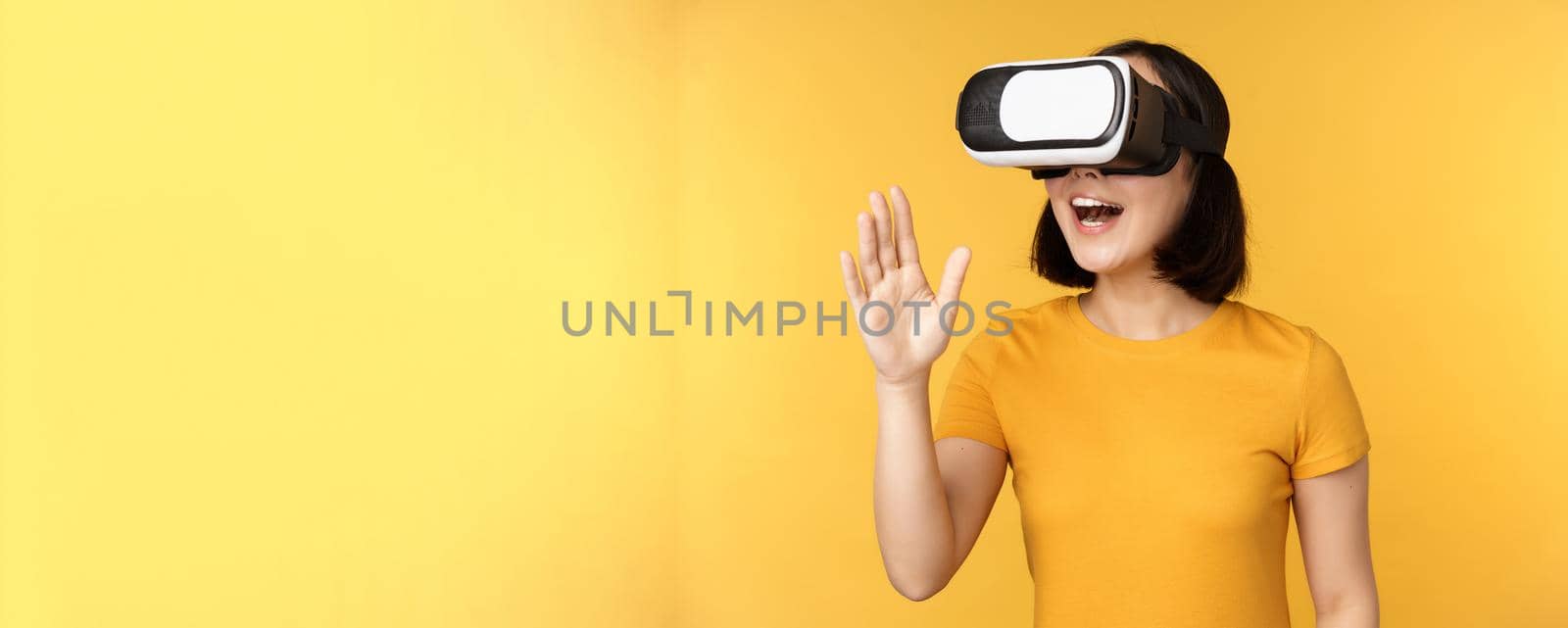 Girl in VR. Beautiful young asian woman, using virtual reality glasses and playing, chatting virtually, standing over yellow background.