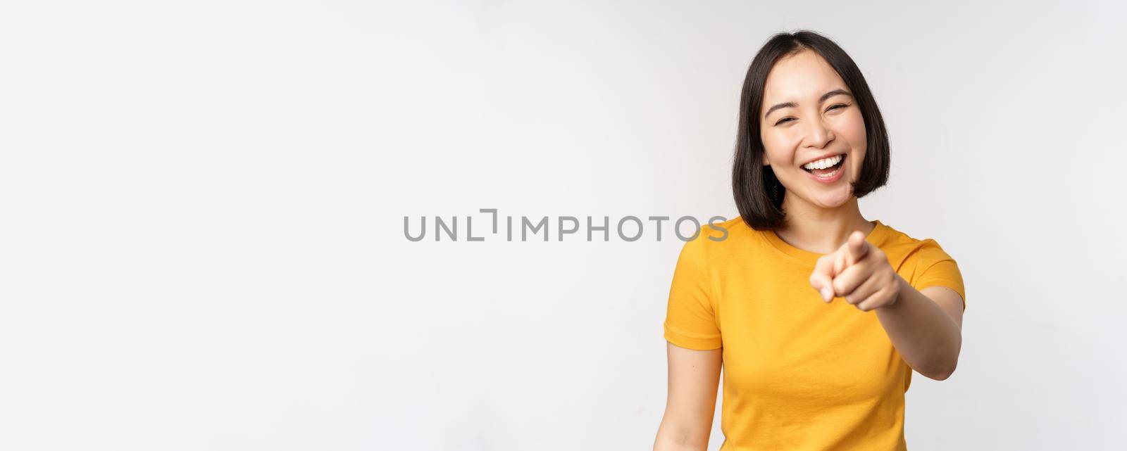 Happy beautiful asian woman laughing, pointing finger at camera and chuckle, smiling carefree, standing in yellow tshirt over white background.