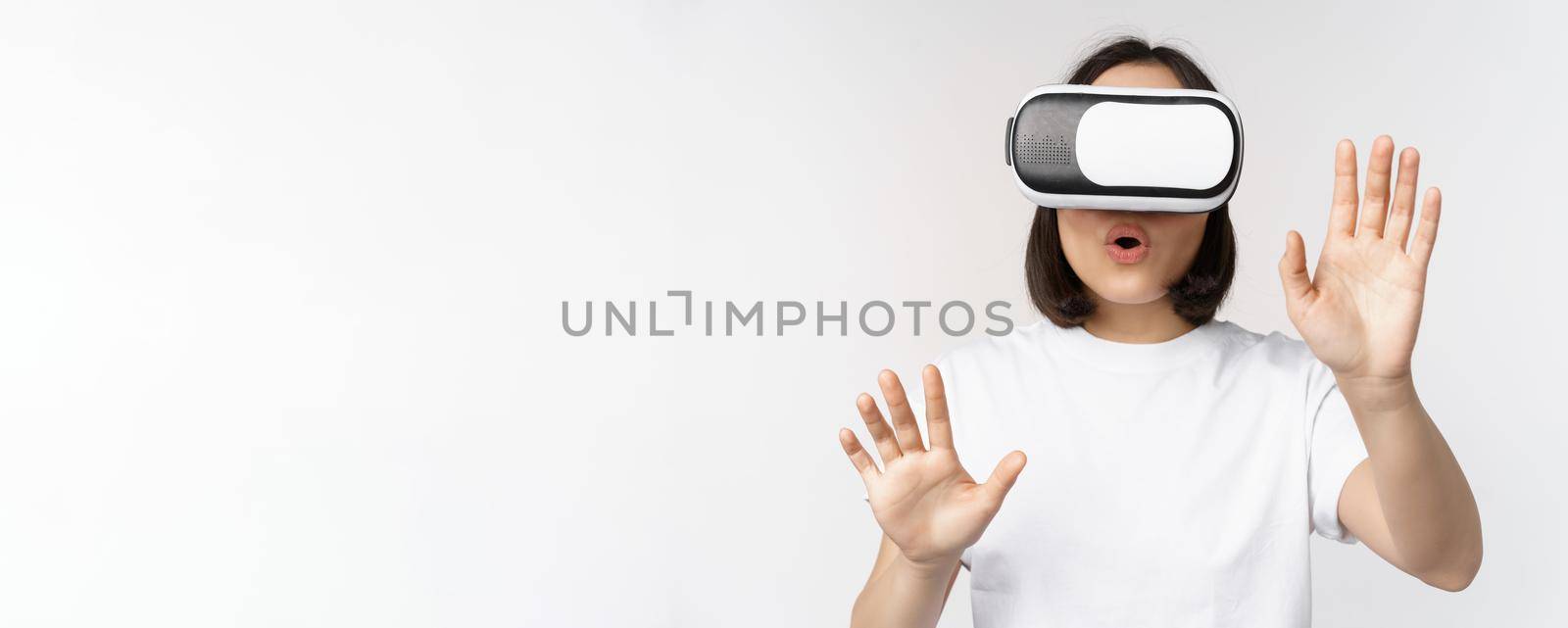 Amused asian girl using VR glasses, virtual reality headset and reaching hands into empty space, touching smth augmented, standing over white background.