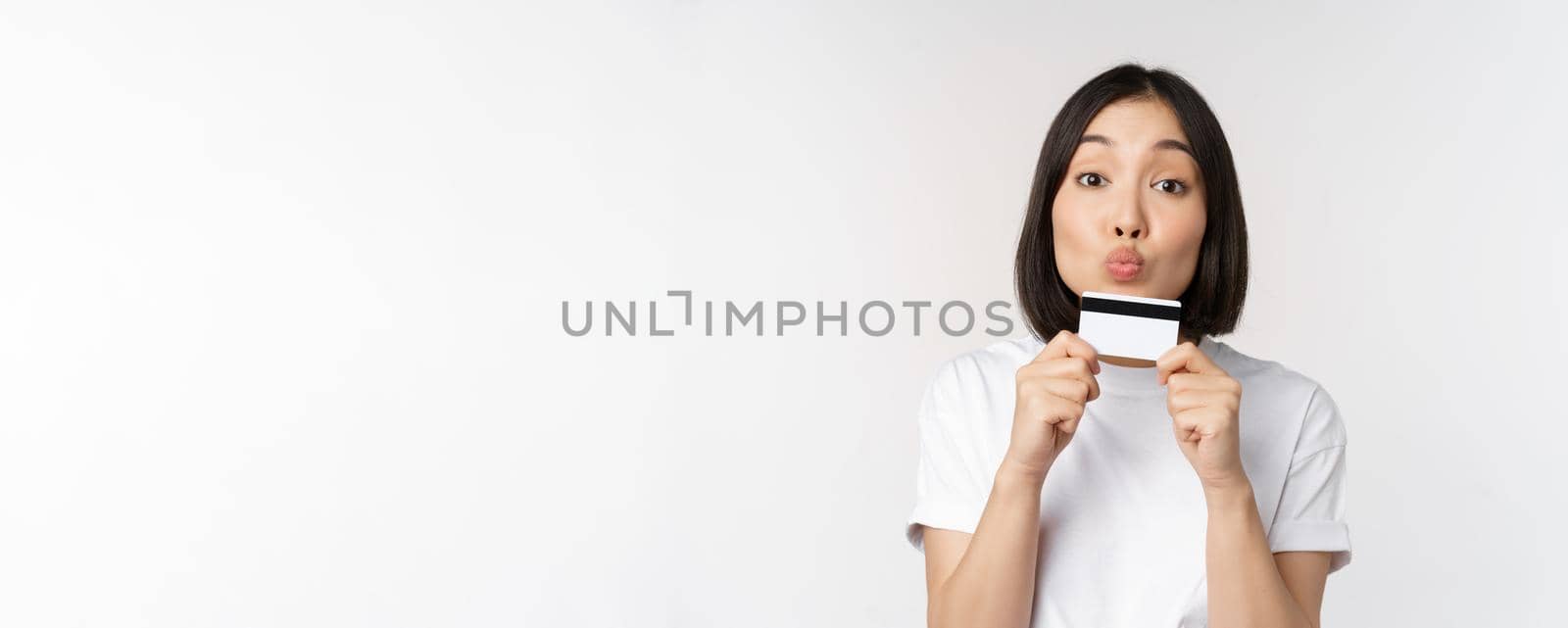 Money and finance concept. Cute japanese girl kissing her credit card, standing in tshirt over white background by Benzoix