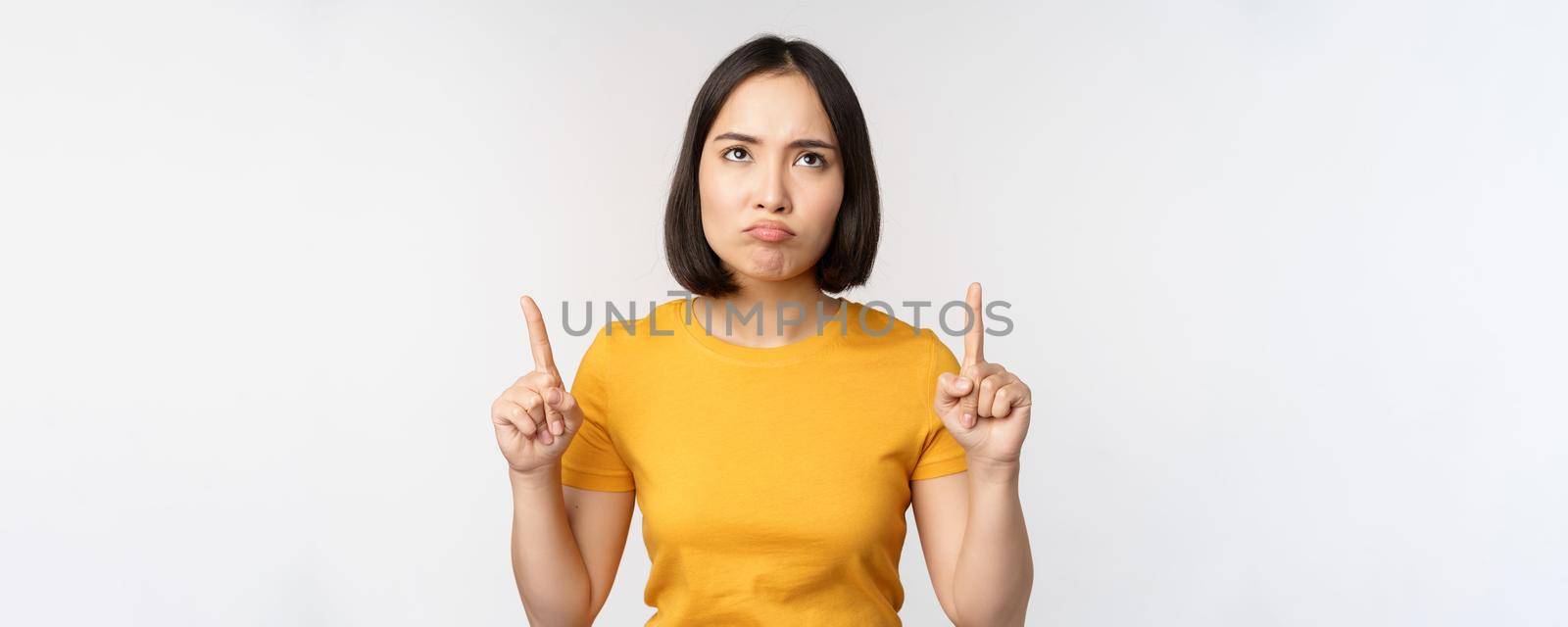 Disappointed asian woman looking, pointing fingers up with angry moody face expression, standing in yellow tshirt over white background by Benzoix